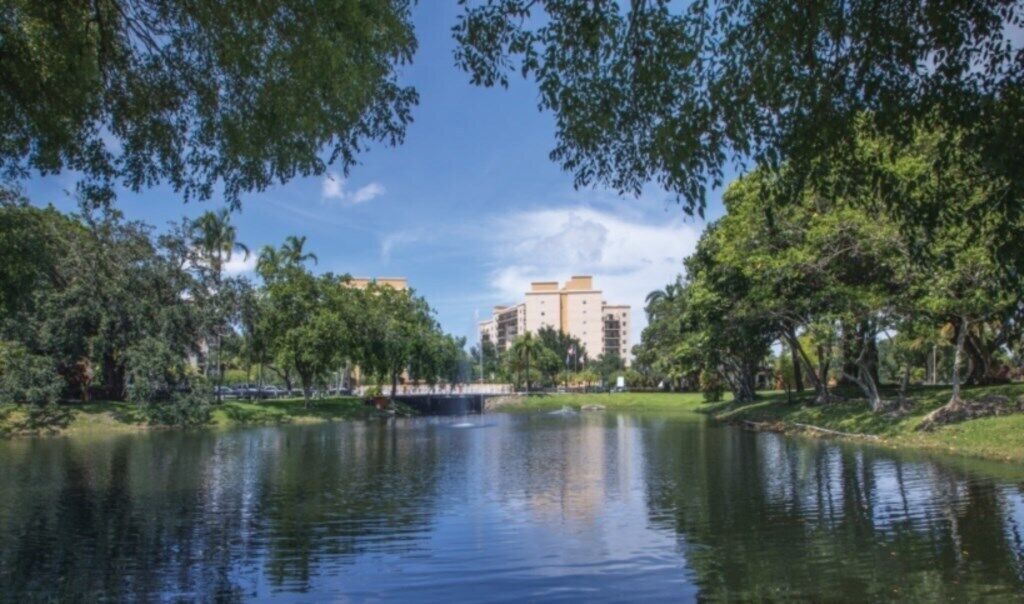 A serene park with a lake, surrounded by lush greenery and trees. In the background, there are multi-story buildings with a modern architectural style.