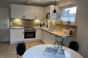 A modern kitchen with white cabinets, a built-in oven, a microwave, and a sink. There is a dining table with chairs and a vase with flowers. - Holiday home in the North Sea with 3 bedrooms (Friedrichskoog-Spitze)