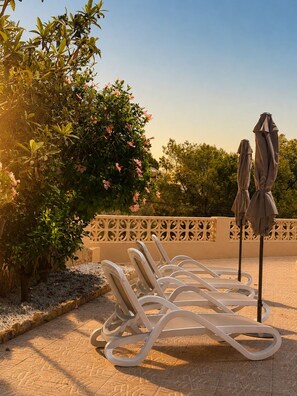 Terraço com espreguiçadeiras e guarda-sóis, rodeado por vegetação e um cercado decorativo.