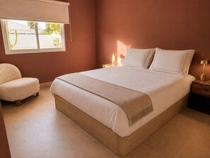 Ein Schlafzimmer mit einem großen Bett, einem Sessel, einem Nachttisch und einem Fenster mit Jalousien.