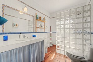 Une salle de bain avec un lavabo blanc, un miroir et des toilettes. On y trouve un porte-serviettes bleu et une étagère avec des articles de toilette.