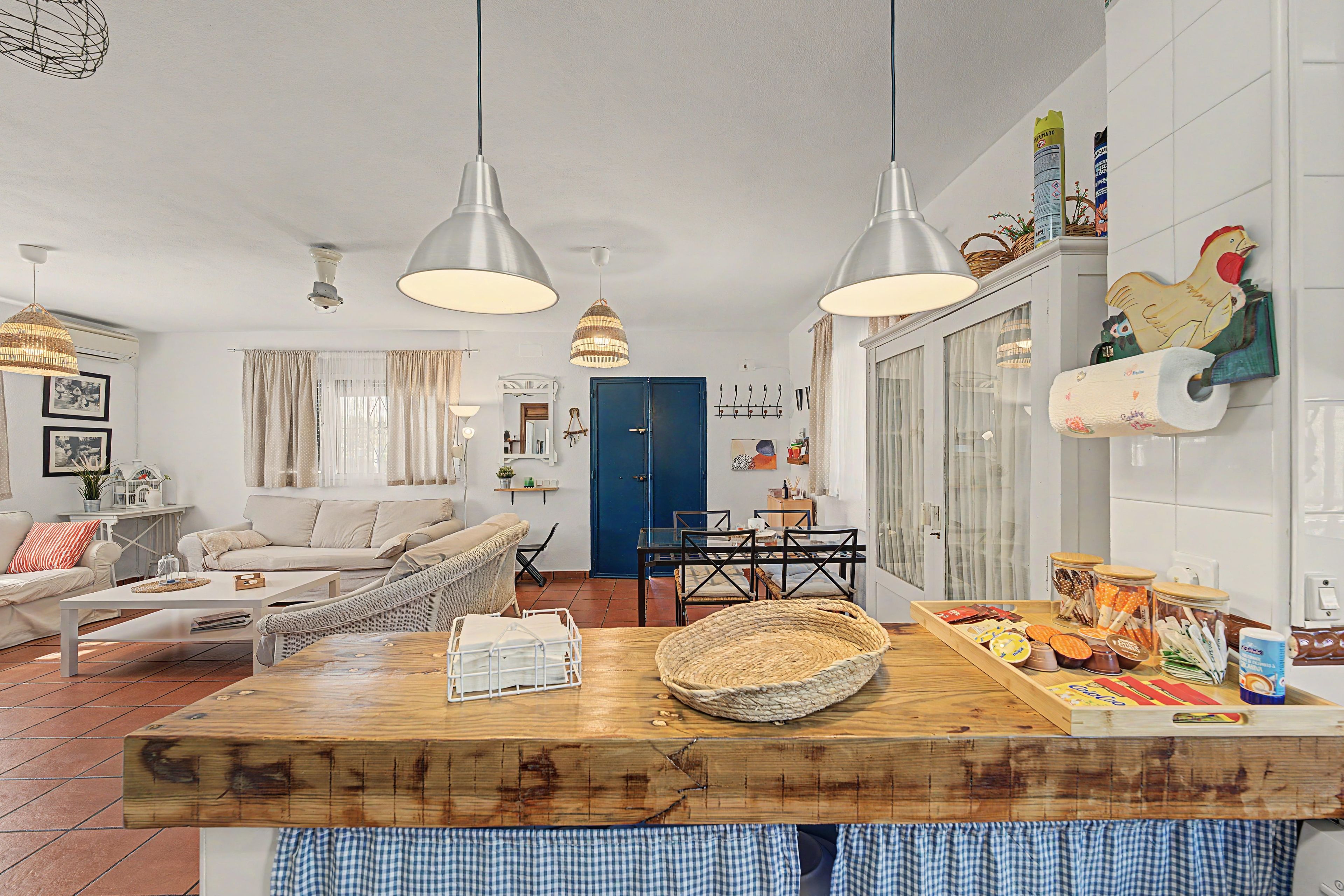 Un salón luminoso con una mesa de comedor de madera, un sofá y una zona de cocina con una lámpara colgante.