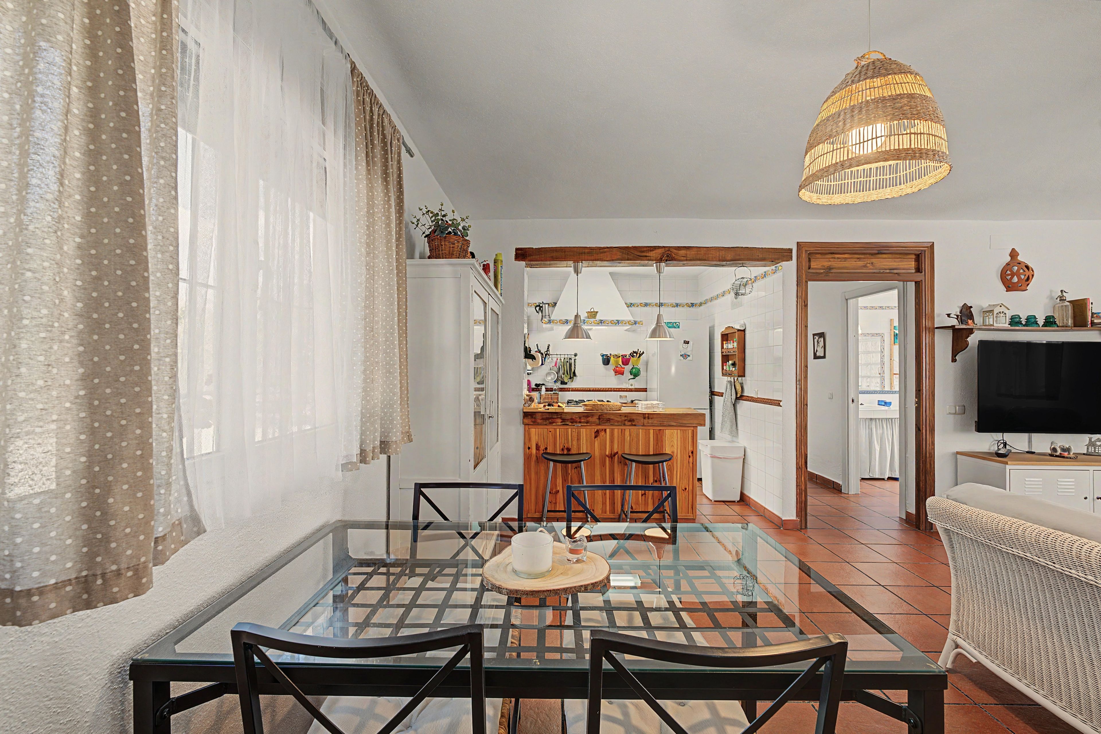 A dining area with a glass table and chairs, a kitchen in the background, and a television on a stand.