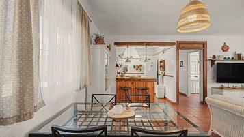 A dining area with a glass table and chairs, a kitchen in the background, and a television on a stand.