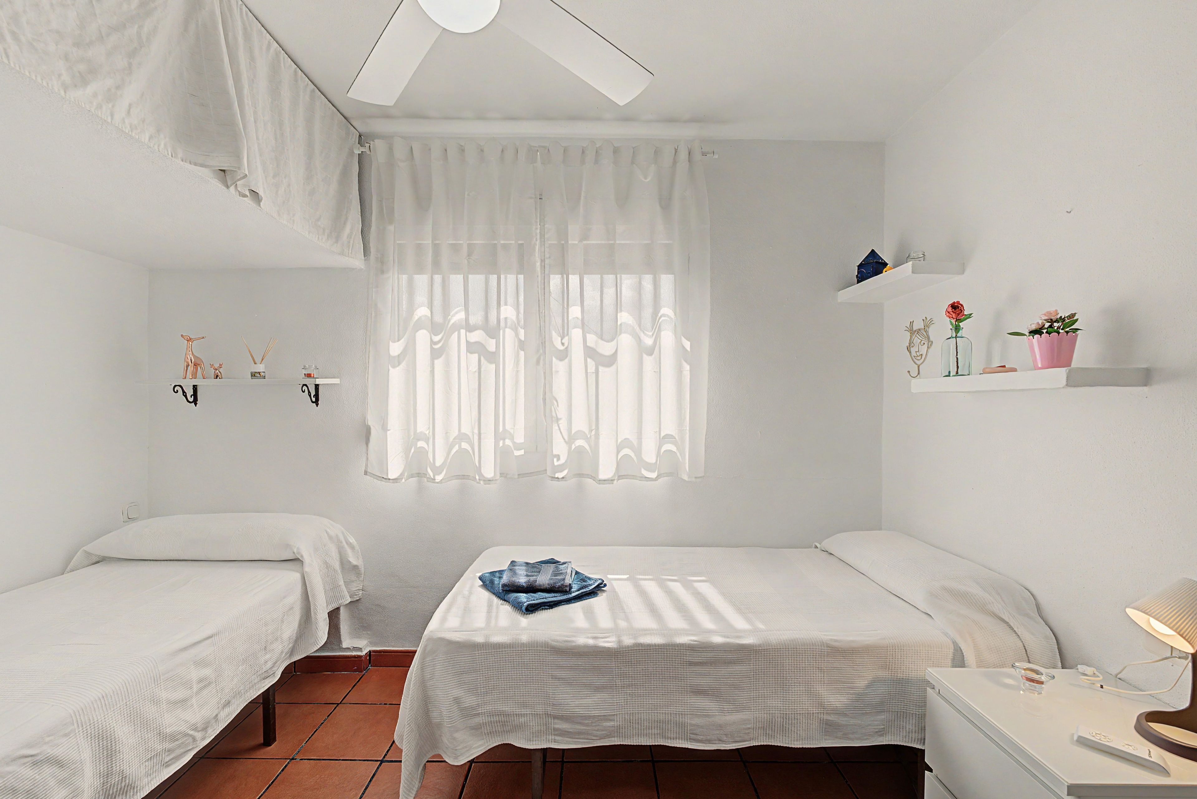 Una habitación con dos camas individuales, una mesita con un objeto azul, un ventilador de techo y una ventana con cortinas translúcidas.