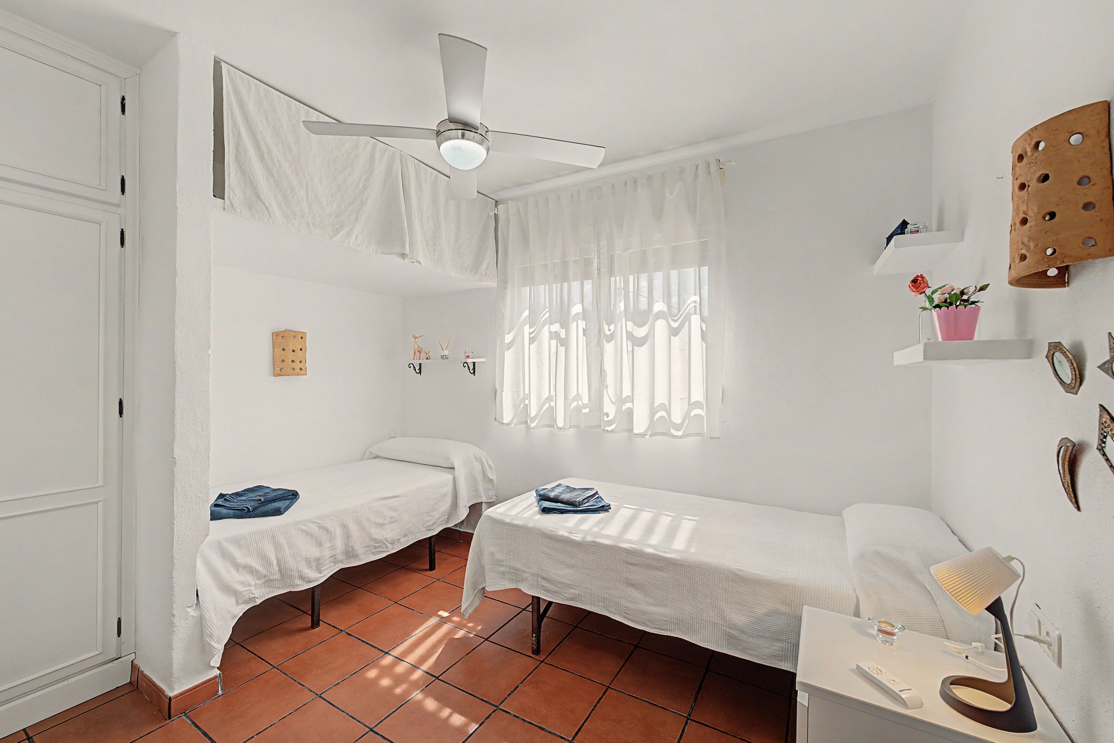 A room with two single beds, a ceiling fan, a window with sheer curtains, and a small shelf with decorative items.