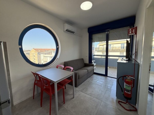 Een ruimtelijk ingerichte leefruimte met een rond raam, een bank, een eettafel met stoelen en een brandblusser.