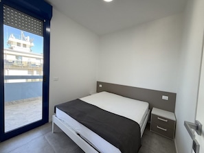 Een slaapkamer met een bed, nachtkastje, raam met jaloezieën en uitzicht op een gebouw.