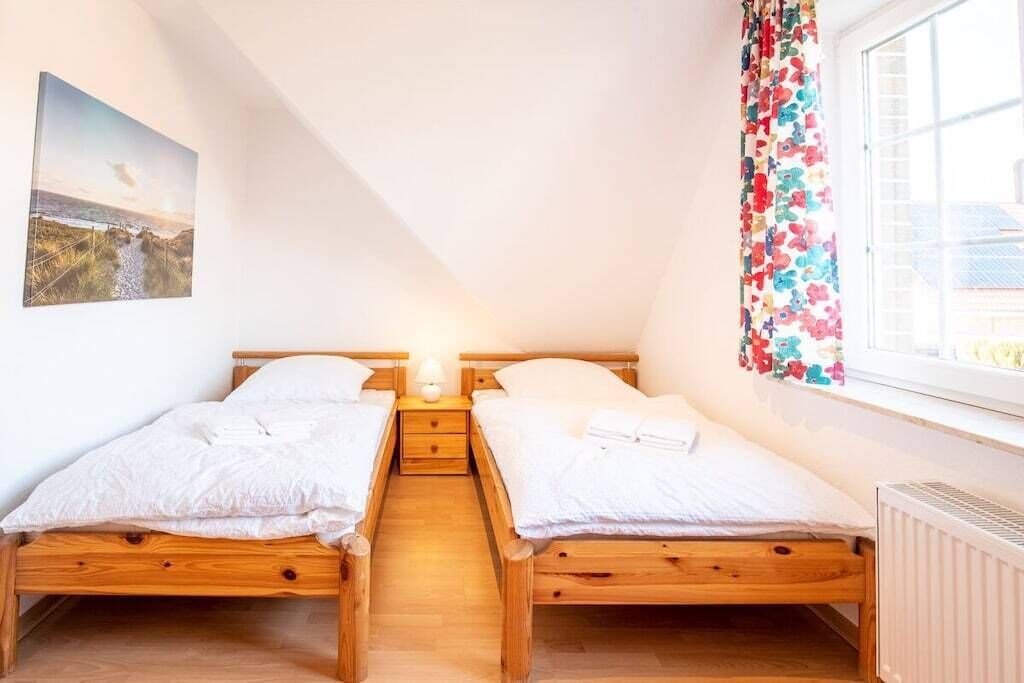 A room with two wooden beds, a nightstand with a lamp, a window with a floral curtain, and a beach scene painting on the wall.