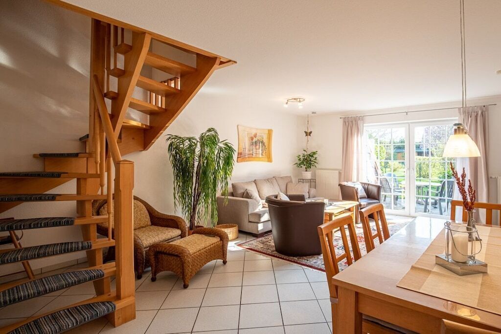 A spacious living area with a wooden staircase, a sofa set, a dining table, and a large window with a view.