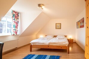 Een slaapkamer met een schuine kap, een bed met witte lakens, een houten vloer, een radiator, een raam met bloemen gordijnen en een klein nachtkastje met een lamp.