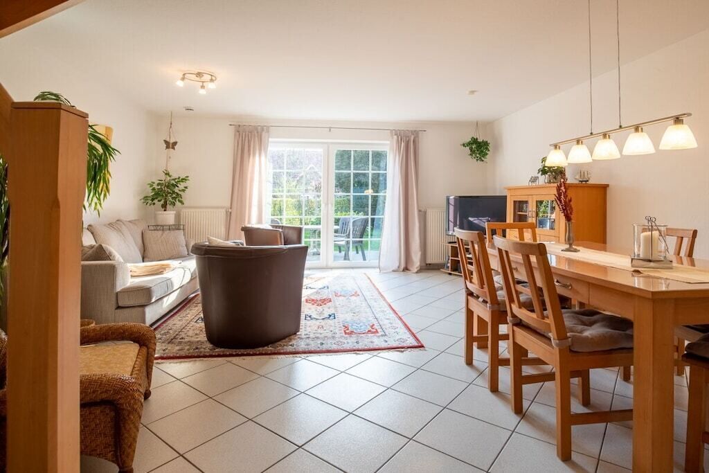 Ein großzügiger Wohnbereich mit Sofa, Couchtisch und Esstisch mit Stühlen. Es gibt ein großes Fenster mit Vorhängen, eine Heizung und einen Fernseher auf einem Schrank.