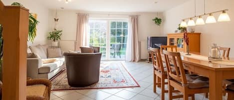 Amplia sala de estar con sofá, una mesa de centro y una mesa de comedor con sillas. Hay un ventanal con cortinas, un radiador y un televisor sobre un mueble.