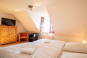 Un dormitorio con techo inclinado, televisor sobre un mueble de madera, una silla, cama con ropa blanca y ventana con cortinas florales.