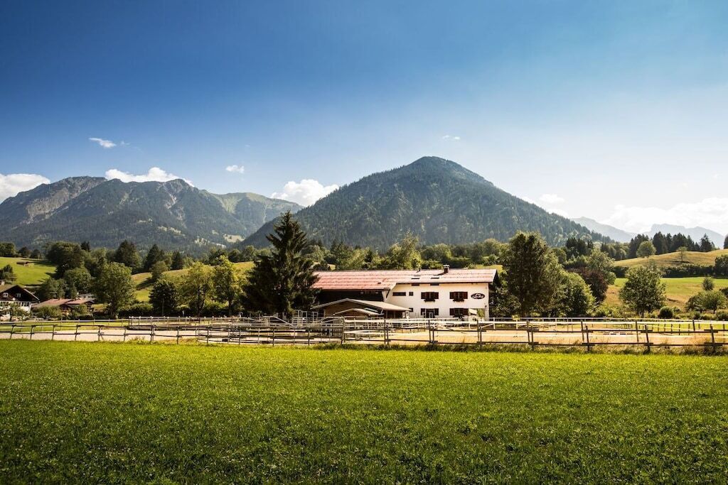 Un paysage rural avec un grand bâtiment, peut-être une ferme ou une maison d’hôtes, entouré de champs verdoyants et avec des montagnes en arrière-plan.