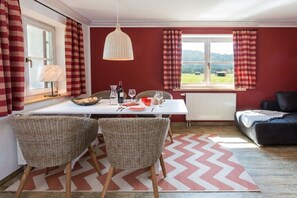 Un coin repas avec une table dressée pour deux personnes, des chaises en osier, un luminaire suspendu et une vue sur l’extérieur à travers une fenêtre ornée de rideaux.