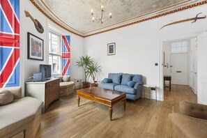 Ein Wohnzimmer mit einem blauen Sofa, einem Holztisch und einem Schreibtisch mit Computer. Zwei Blumentöpfe, eine Chandelier und ein gerahmtes Bild an der Wand sind ebenfalls zu sehen.