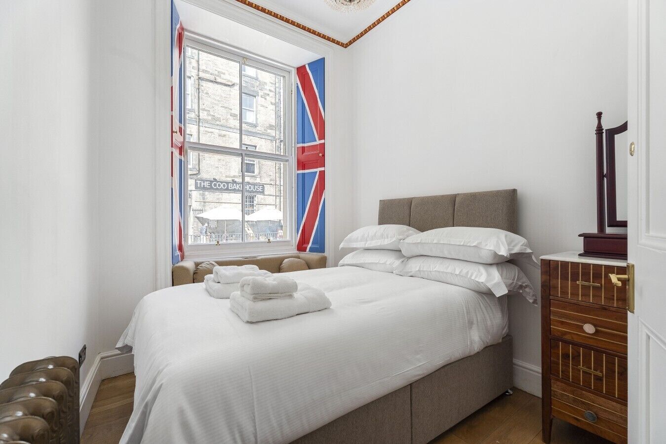 A neatly made bed with white linens and pillows, a wooden dresser with drawers, and a window with a view of a building labeled 'The Coo Bakery House.'