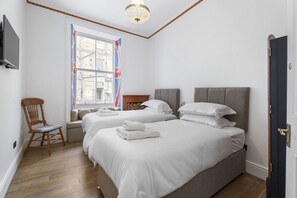 Ein Hotelzimmer mit zwei Betten, einem Holzstuhl, einem Fernseher und einem Fenster mit Blick auf ein Gebäude.