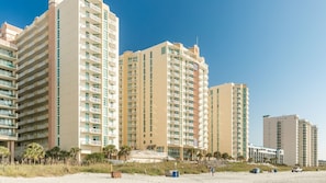 A beachfront hotel with multiple high-rise buildings, a sandy beach, and palm trees. - North Myrtle Beach | Oceanfront Family Stay | Pool & Beach | 3BR Sleeps 10 (North Myrtle Beach)
