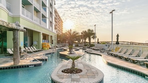 A swimming pool with lounge chairs, a palm tree, and a building with balconies. - North Myrtle Beach | Oceanfront Family Stay | Pool & Beach | 3BR Sleeps 10 (North Myrtle Beach)