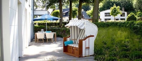 Una terraza de madera con mobiliario exterior, incluyendo una silla de mimbre y una mesa con sillas, rodeada de abundante vegetación y árboles.