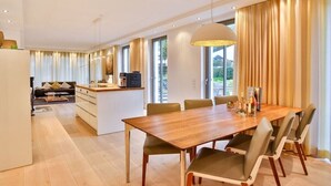 Un comedor moderno con una mesa y sillas de madera, una cocina al fondo y ventanas grandes con cortinas.