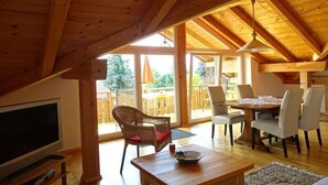 A spacious room with a wooden ceiling, a television, a dining table with chairs, and a large window providing a view of the outdoors. - Holiday apartment with 1 bedroom (Seebruck)