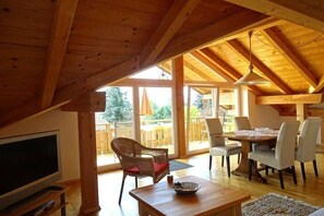 Un'ampia stanza con soffitto in legno, televisione, tavolo da pranzo con sedie e un'ampia finestra che offre vista sull'esterno.