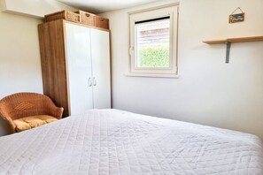 A bedroom with a bed, a wardrobe, a wicker chair, a window, and a shelf. - Holiday apartment OK16 Oostkapelle (Oostkapelle)