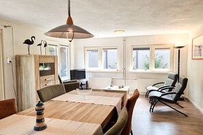 A dining area with a wooden table, chairs, a hanging light fixture, and a view of the outdoors through windows. - Holiday apartment OK16 Oostkapelle (Oostkapelle)