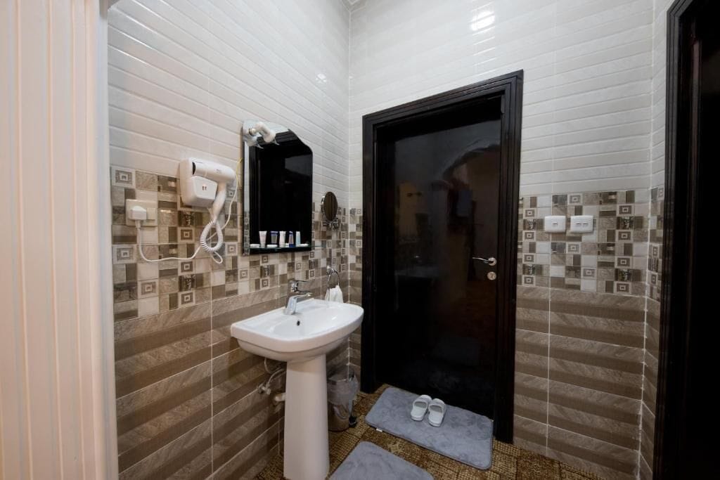 Une salle de bain avec un lavabo blanc, un sèche-cheveux et un miroir. On y trouve une porte avec un cadre foncé et un mur recouvert de carreaux formant un motif.
