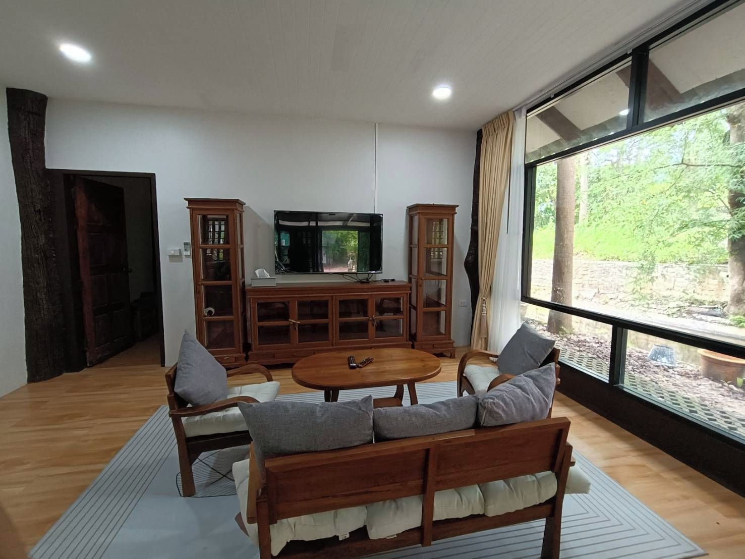 Ett vardagsrum med en soffa i trä, ett soffbord och en tv som står på ett TV-bord. Det finns två skåp i trä och ett stort fönster med utsikt över grönområden.