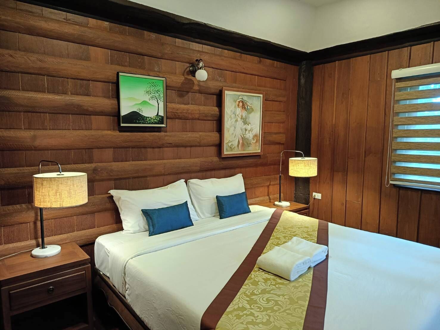 Quarto de hotel com parede revestida de madeira, cama com roupa de cama branca e azul, duas luminárias de cabeceira e obras de arte.