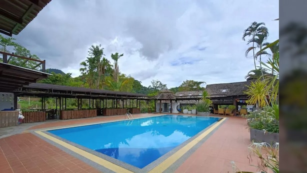 A rectangular swimming pool with a tiled deck surrounding it, a covered seating area to the left, and a building with a thatched roof to the right.