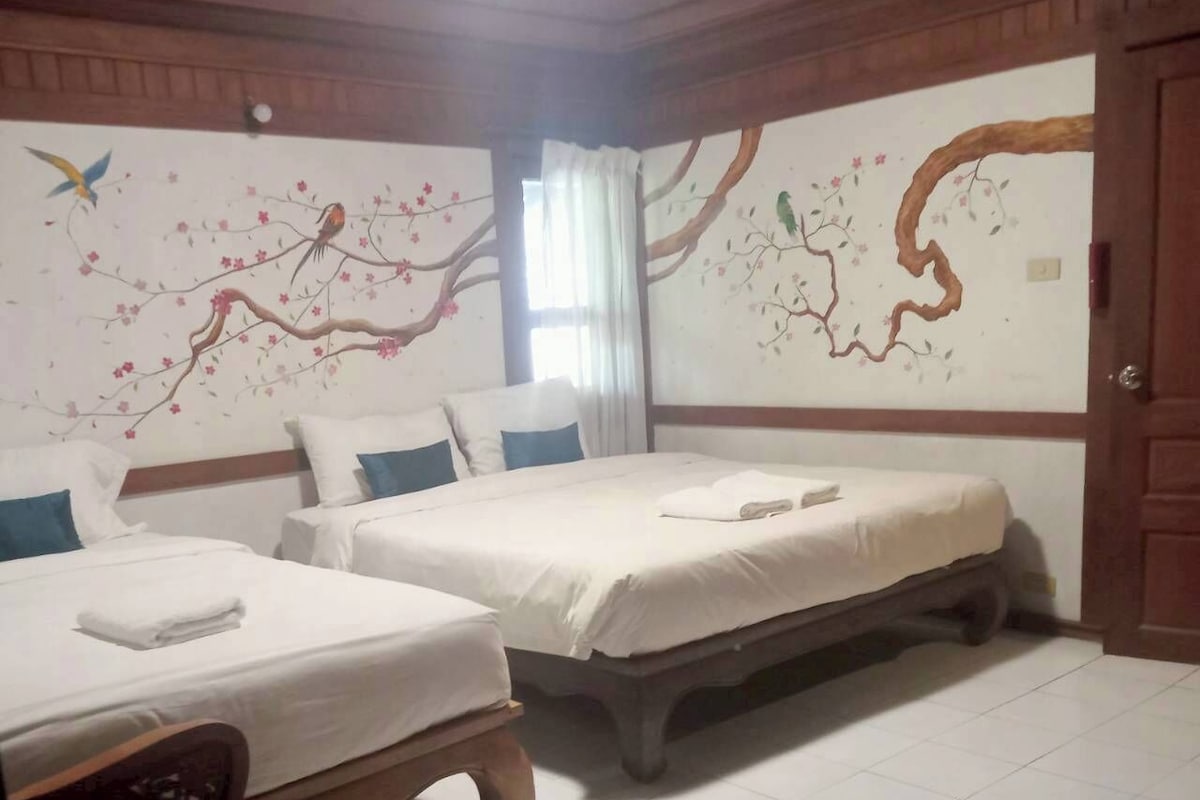 Habitación de hotel con dos camas, una silla de madera y un mural en la pared con un árbol y un pájaro.