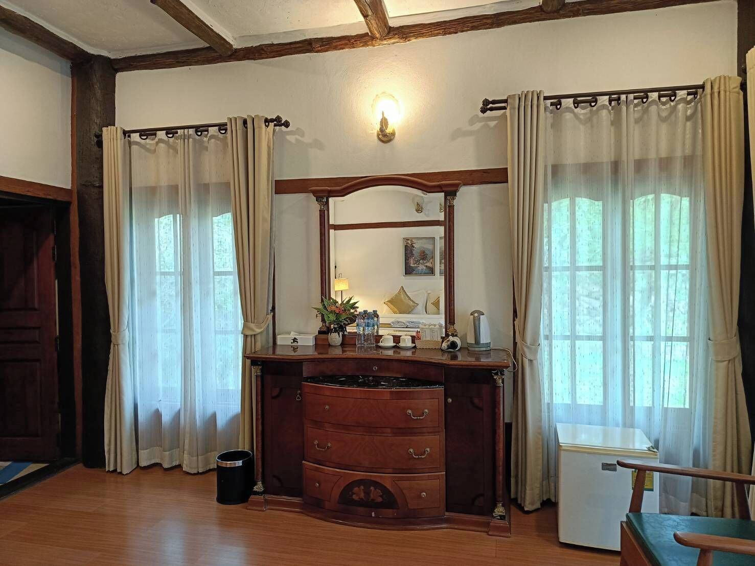 A room with a wooden dresser, a mirror, a small table with a lamp, and a window with curtains.