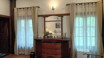 A room with a wooden dresser, a mirror, a small table with a lamp, and a window with curtains.