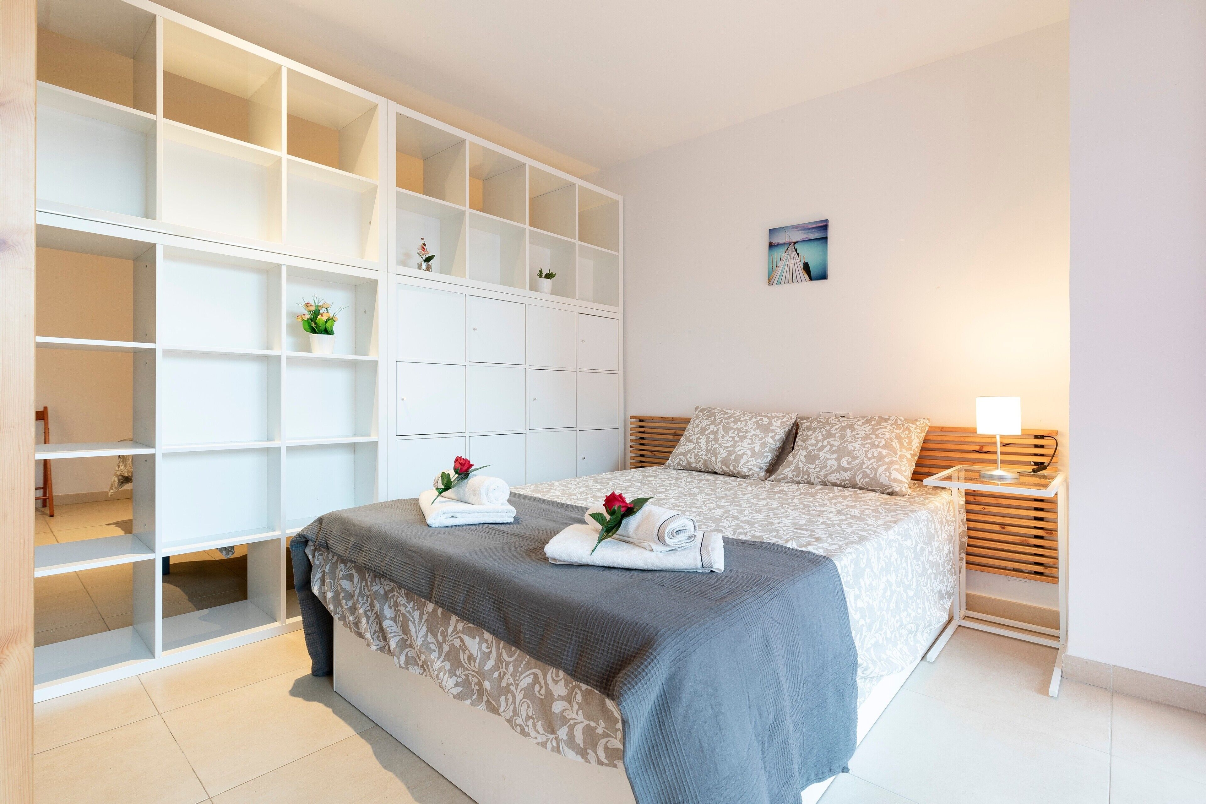 A bedroom with a bed, a nightstand, a lamp, a wardrobe with multiple shelves, and a small picture on the wall.