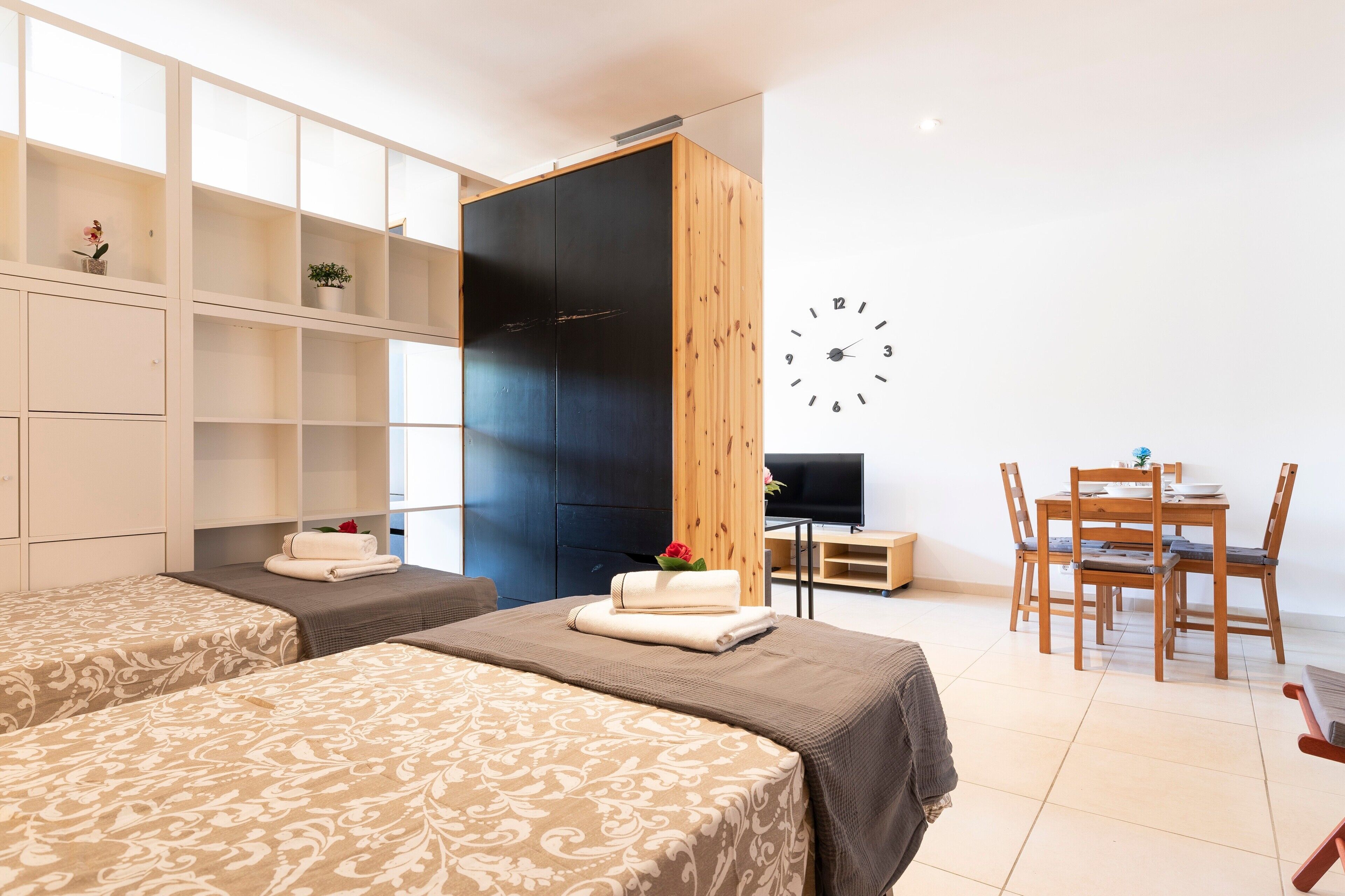 A hotel room with two beds, a wardrobe, a desk with a TV, and a dining area.