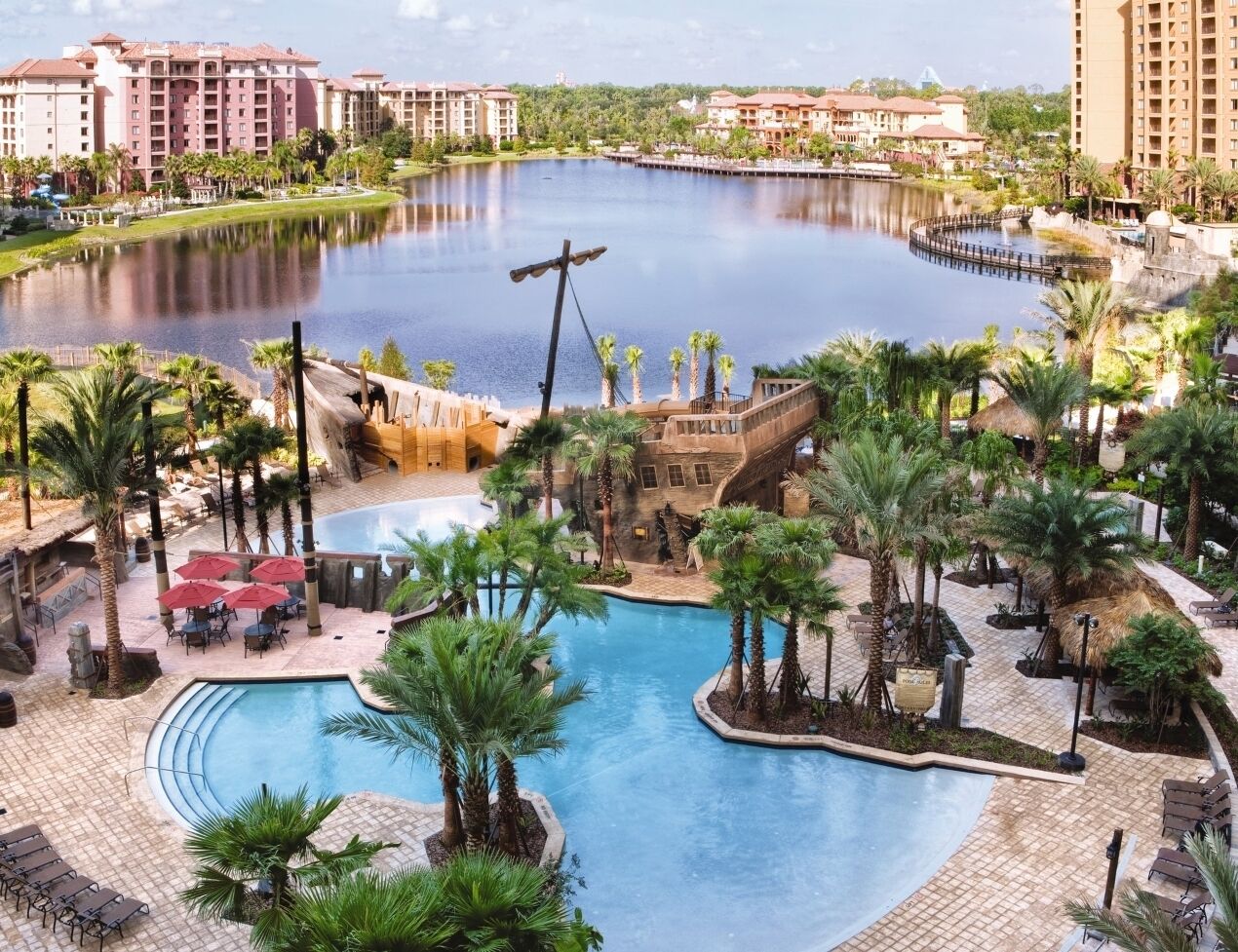 A resort with a central pool, palm trees, and a water body.