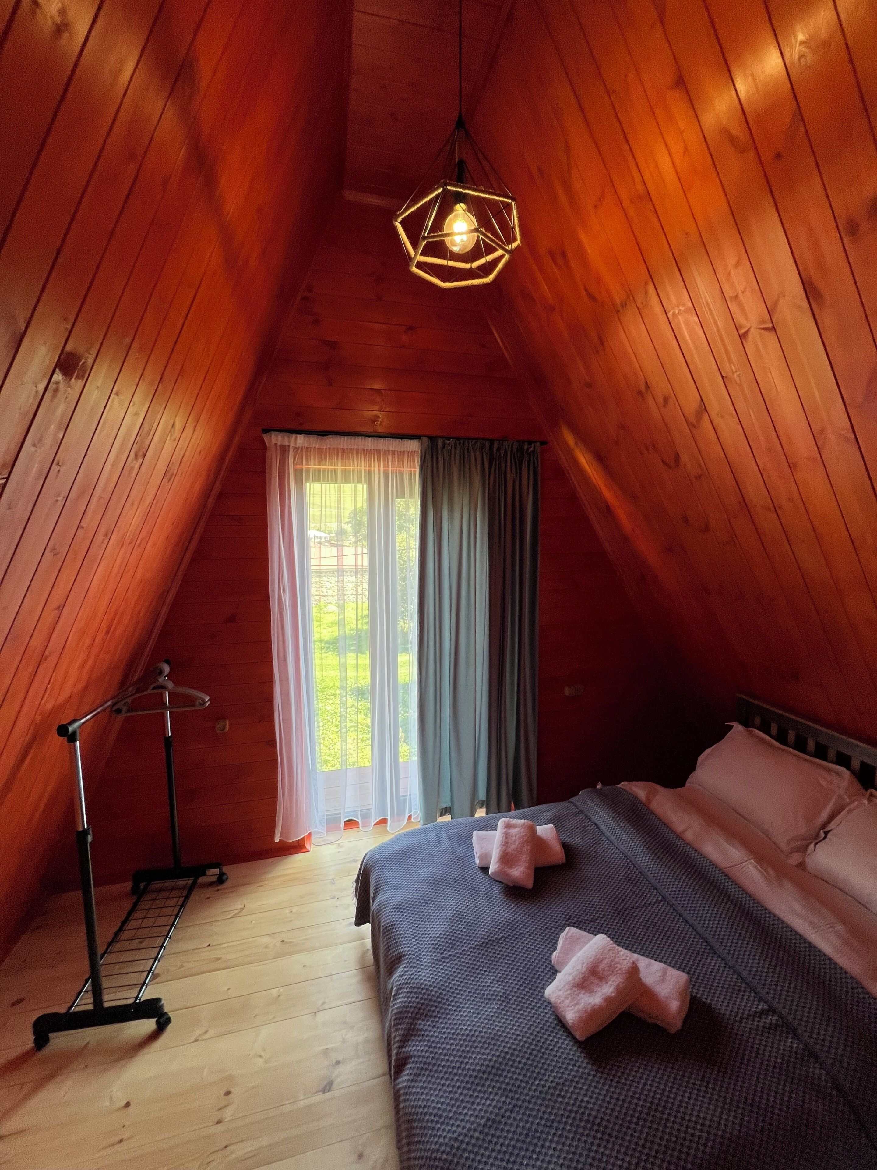 A room with a sloped ceiling, a bed with pillows, a hanging light fixture, and a window with curtains.