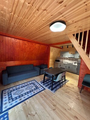 A cozy room with a wooden ceiling, a sofa, a dining table with chairs, and a kitchen area visible through an open space. - Perfect 2-bedroom cottage in charming Achkhoti for a relaxing stay (Achkhoti)