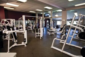 Uma academia com diversos equipamentos de exercícios, uma tela de televisão e uma grande janela ao fundo.