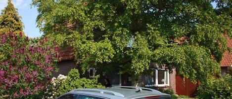 A silver car parked in front of a house with a white picket fence, surrounded by lush greenery and flowering plants.