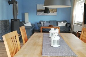 Een woonkamer met een bank, een salontafel en een eettafel met stoelen. Er is een open haard met schoorsteen en een lamp boven de bank.