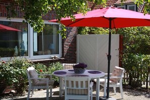 Een terras met een tafel en stoelen, een rode parasol en een witte schutting.