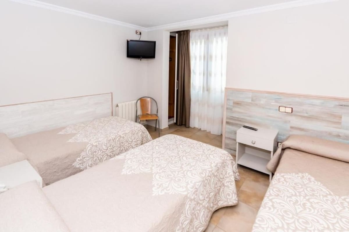 A hotel room with two beds, a TV mounted on the wall, a chair, a small table, and a window with curtains.
