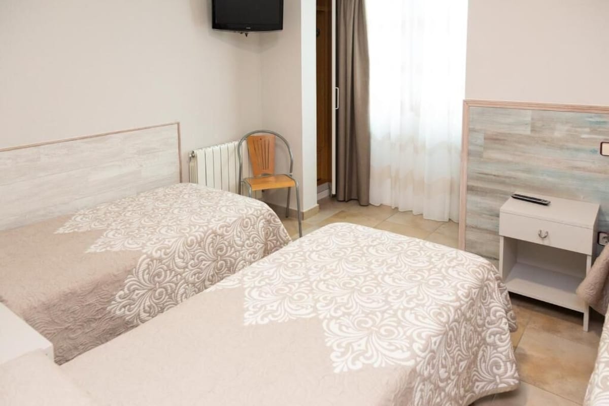 A hotel room with two beds, a chair, a TV, a nightstand, and a window with curtains.