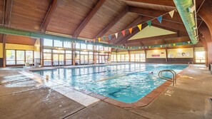 An indoor swimming pool with clear lanes, wooden beams, and a view of the outside through large windows.